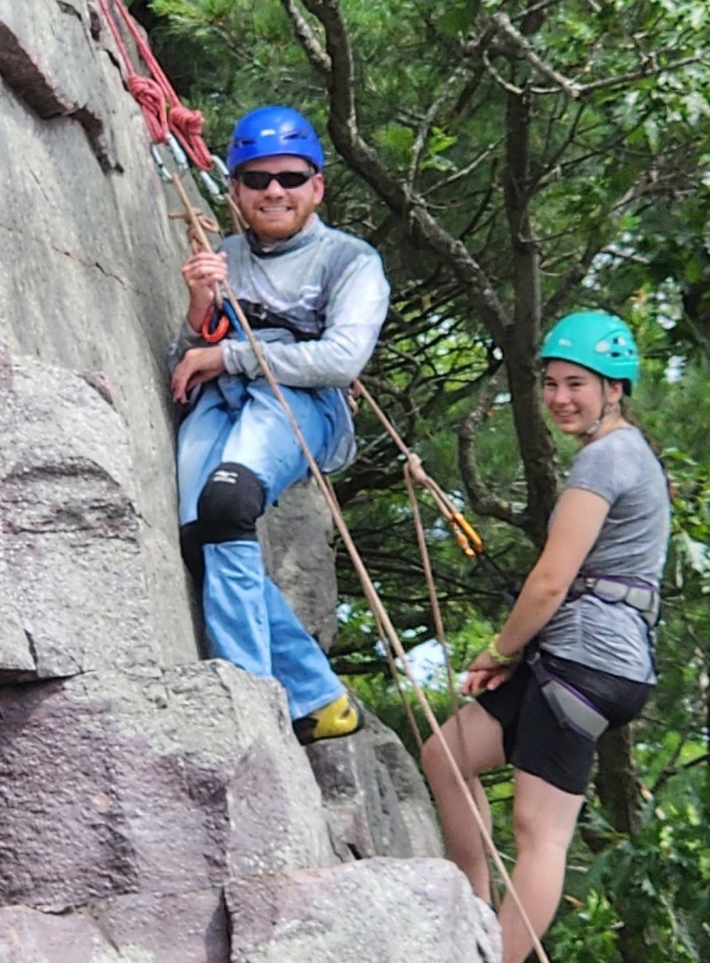 A young man wearing a blue helmet, sunglasses, and long-sleeve shirt, secured to a rock face with a harness and ropes. A young woman wearing a green helmet and climbing harness stands beside him on the ledge.