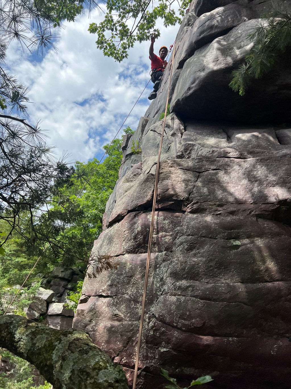 A young man with determination on his face signaling toward someone below. He is wearing a red shirt, a yellow helmet, and a full-body harness system.